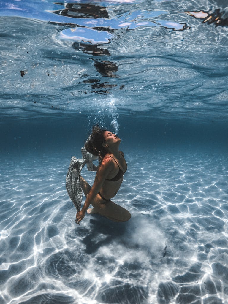 A woman gracefully swims underwater, exhaling bubbles in the clear ocean.